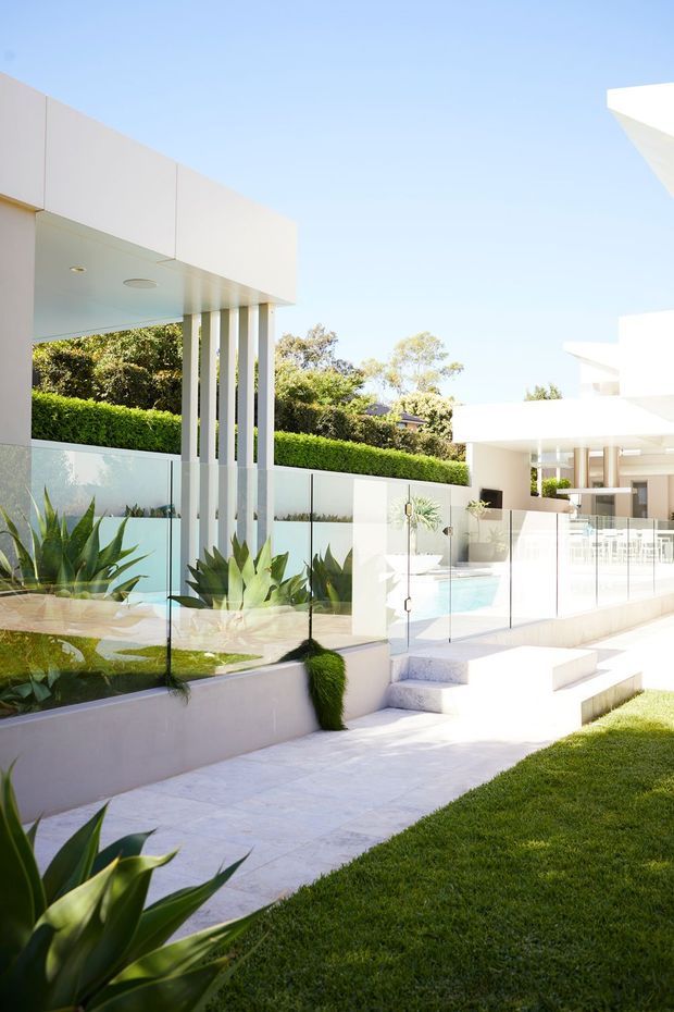 Bright exterior view of a sophisticated modern residence with crisp white cubic forms, deep roof overhangs, slender vertical columns, extensive glass balustrades, landscaped succulent planting, a paved walkway and steps, and a swimming pool terrace. The scene has a clean, minimalist architectural character in strong sunlight.