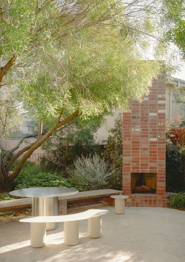 A landscaped outdoor courtyard or patio with a built-in red brick fireplace, pale paved flooring, curved built-in and freestanding seating, and a round metal table. Mature leafy planting creates a shaded, garden-like setting, while the brick fireplace and integrated bench define the outdoor entertaining area.