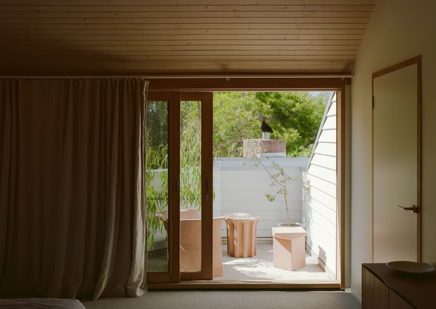 A warm, minimalist interior looking through wide timber-framed glazed sliding doors onto a small private terrace or courtyard. The room has a pale timber slatted ceiling, dark full-height curtains, neutral walls, a low dark timber cabinet, and a simple table lamp. Outside, the terrace includes pale paving, sculptural light-coloured furniture, a rendered privacy wall, and a white horizontal-clad exterior wall.