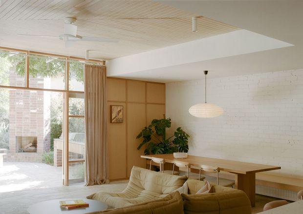 Bright, warm-toned open-plan living and dining area with mid-century influences. The room features a large glazed opening to an outdoor courtyard, timber-framed glazing, a timber slatted ceiling, white painted brick walls, built-in timber cabinetry, a long timber dining table, pendant lighting, and a cream sectional sofa. Indoor plants and natural materials create an airy, relaxed atmosphere.