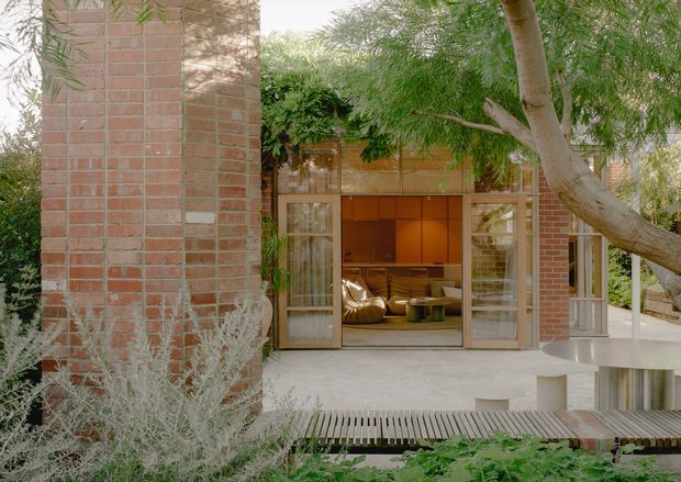 Exterior view of a brick-clad residence opening onto a landscaped courtyard or garden. Large timber-framed glazed doors are open to a warm-toned living area, with a pale concrete terrace, timber slatted walkway, mature trees and planting in the foreground. The composition highlights the indoor-outdoor connection and substantial brick wall construction.