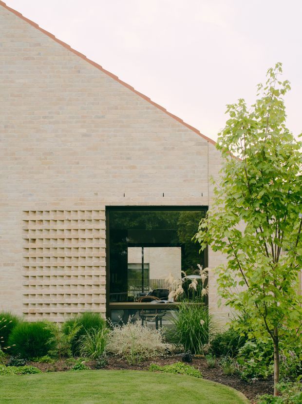 Exterior view of a pale beige brick building with a steeply sloping gable wall and terracotta-edged roof. A large black-framed glazed opening provides a view into the interior, while a textured projecting brick screen sits beside it. The facade is integrated into a landscaped garden with ornamental grasses, shrubs, a small tree, and lawn.