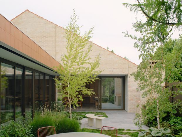 A landscaped courtyard view of a contemporary low-rise building with pale brick gabled volumes, dark-framed floor-to-ceiling glazing, and a warm copper-toned metal-clad wing. Young trees, grasses, planting beds, paved paths, and outdoor furniture create a sheltered garden setting.