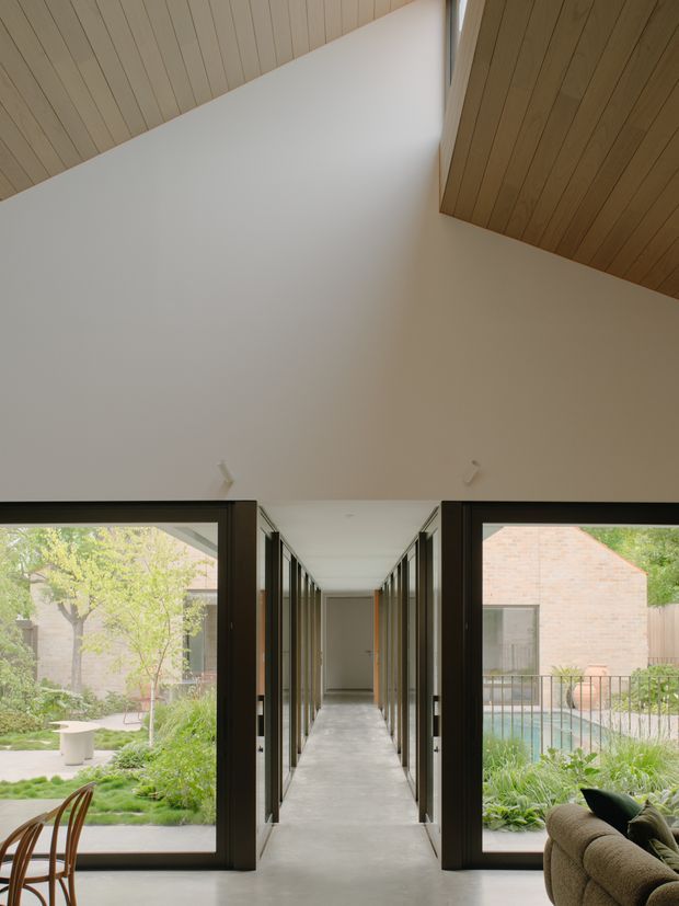 Interior view looking down a long, gallery-like hallway with smooth pale concrete flooring, dark-framed full-height glazed doors on both sides, and a dramatic high angled white ceiling intersected by warm timber-lined ceiling planes. Landscaped courtyards and adjacent brick structures are visible through the glazing, while part of a lounge chair and side table appear in the foreground.