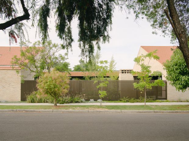 Street-facing view of a low-rise residential property behind a landscaped front boundary. Pale brick walls, terracotta tiled pitched roofs, and a tall dark timber or metal privacy fence and gate are partially screened by mature trees and planting. A footpath and quiet roadway occupy the foreground.