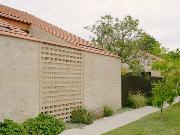 Exterior view of a low-rise masonry building with pale rendered or stone-like brick walls, a terracotta tiled pitched roof, and a prominent breeze-block screen panel. A narrow concrete walkway runs alongside landscaped garden planting and a dark boundary fence. The image has a soft, understated residential architectural character.
