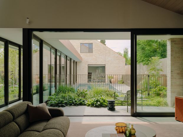 A contemporary living room opening through expansive black-framed glazing onto a planted courtyard. The room features a low curved dark sofa, a round coffee table, pale walls, and a timber-lined ceiling, with a brick courtyard building and lush garden visible beyond.