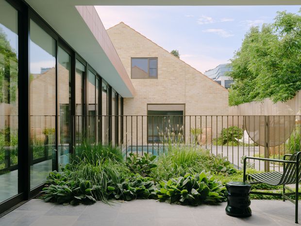 A landscaped courtyard terrace viewed from a modern glazed building. Full-height black-framed windows line the left side, opening onto a paved outdoor area with dense ornamental planting, a slender metal balustrade, and black outdoor seating. Beyond the courtyard is a simple pale brick gabled building with large windows, while trees and a contemporary urban building are visible in the background.
