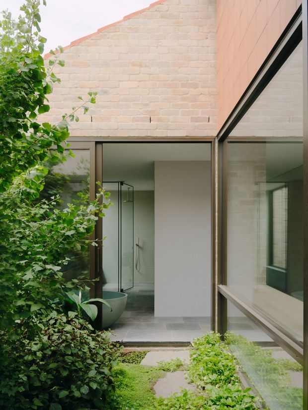 A view into a compact landscaped courtyard framed by exposed pale brick walls and dark-framed floor-to-ceiling glazing. The courtyard contains dense green planting, mossy groundcover, stepping stones, and a partially visible contemporary bathroom with a freestanding rounded bathtub, glass shower enclosure, and neutral grey floor tiles. The composition highlights a strong indoor-outdoor connection and reflective glazing.