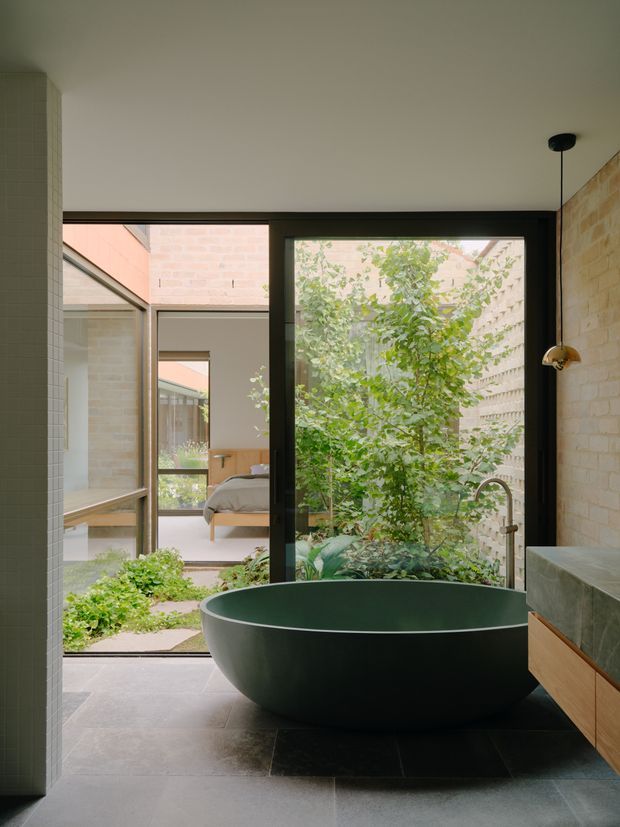 A contemporary bathroom with a freestanding matte dark green bathtub positioned beside large black-framed glazing overlooking a leafy internal courtyard. Pale exposed brick walls, grey tiled flooring, a timber-fronted vanity, and a sculptural brass pendant create a calm, natural atmosphere. A bedroom is visible through the open glazed doorway beyond the courtyard.