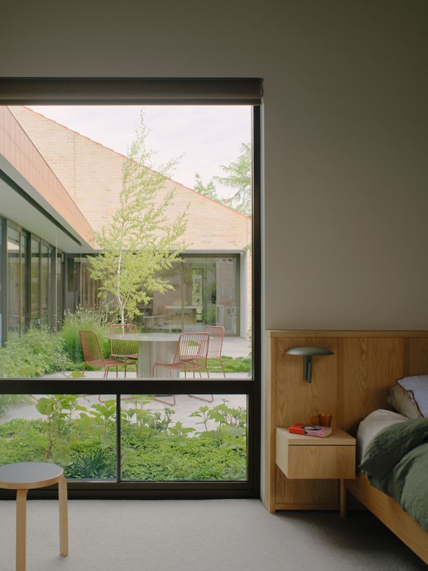 A contemporary bedroom with a large dark-framed picture window overlooking a landscaped courtyard. The room features a low timber bed with an integrated timber headboard and bedside storage, a compact wall-mounted reading light, neutral carpet, and a small round side table. Through the glazing, a planted courtyard with outdoor seating and a brick-clad building is visible.
