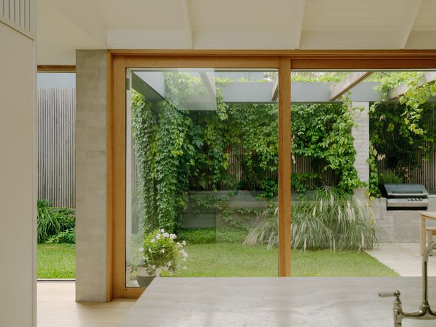 A bright kitchen interior viewed toward a lush courtyard through large timber-framed glazed doors. The foreground shows a pale stone kitchen benchtop with a traditional metal tap, while outside is a landscaped garden with climbing greenery, ornamental grasses, a paved outdoor cooking area and barbecue. The room has a white-painted timber-lined ceiling, pale concrete-toned walls and warm timber framing.