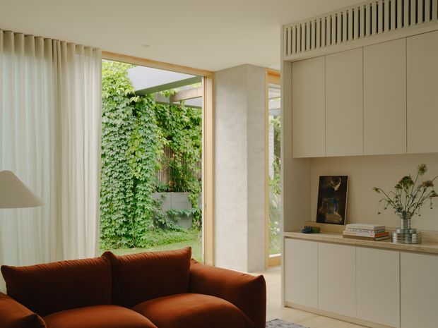 Warm contemporary living area with a deep rust-brown upholstered sofa in the foreground, full-height cream curtains, pale neutral walls, and bespoke light cabinetry along the right wall. A large glazed opening connects the room to a lush private courtyard garden, while a high slatted ventilation or clerestory detail adds architectural interest above the cabinetry.