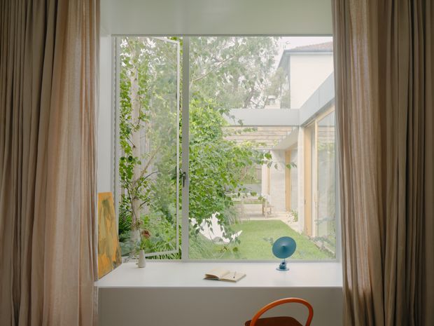 A softly lit interior view looking through a large floor-to-ceiling window into a leafy courtyard. Full-height beige curtains frame the opening, with a deep white window seat or sill holding a small plant, artwork, book and blue decorative object. A glimpse of a contemporary courtyard building with glazed connections and a covered walkway is visible beyond.