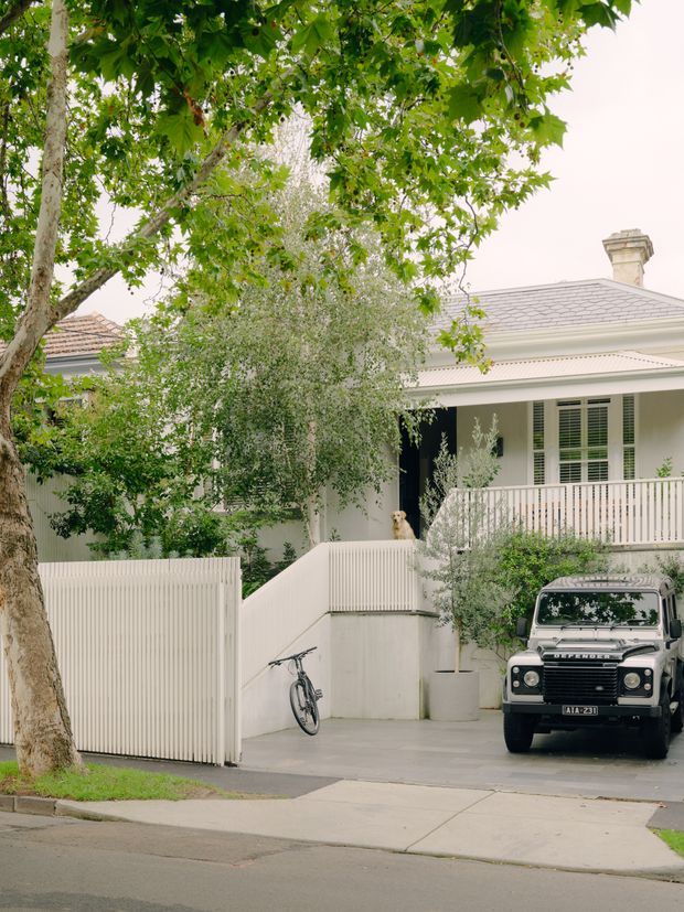 Street-facing view of a refined white residential facade, likely a traditional or heritage-style home. The property features a pitched grey tiled roof, decorative eaves and cornice detailing, a chimney, white rendered walls, shuttered windows, a raised balcony/verandah with vertical balustrading, and a gated driveway. Mature trees and planting partially frame the house, while a parked vehicle is visible in the driveway.