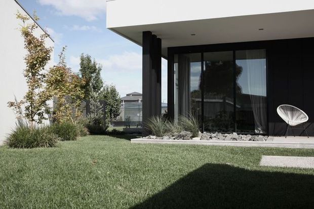 Modern single-storey home exterior with a crisp white flat roof overhang, dark vertical wall cladding, and expansive floor-to-ceiling glazing. A minimalist landscaped garden with ornamental grasses, pale gravel, a lawn, concrete stepping slabs, and a white sculptural outdoor chair frames the covered patio.
