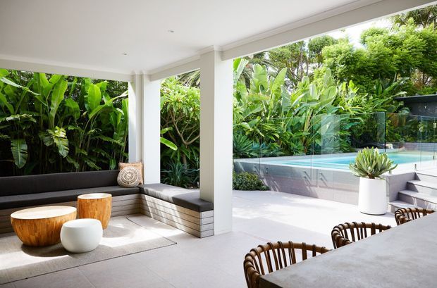 Bright contemporary indoor-outdoor living and dining area with floor-to-ceiling glazing opening onto a lush tropical garden and swimming pool. The space features a built-in dark upholstered sectional, light tiled flooring, sculptural wooden coffee tables, a pale dining table with woven chairs, and a covered terrace-like connection to the pool.