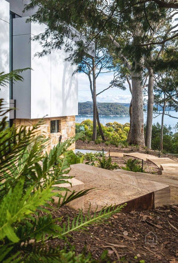 A contemporary multi-level home set into a landscaped, wooded slope overlooking a calm coastal inlet. The visible facade combines crisp white panel cladding and extensive dark-framed glazing with a lower section of warm, irregular natural stone. Broad stone or concrete terraces and steps weave through native planting and mature trees, with water and a wooded shoreline visible beyond.