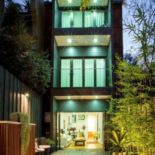 A narrow multi-storey townhouse facade photographed at dusk, with dark vertical cladding, stacked balconies or recessed window bays, extensive glazed openings, and integrated warm exterior lighting. Lush planted screening and a brightly illuminated ground-floor living area create a sheltered urban courtyard-like frontage.