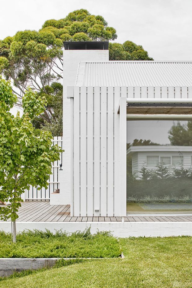 Exterior view of a white, vertically clad residential building with a corrugated metal pitched roof, chimney-like upper structure, large glazed opening, and timber decking. Mature landscaping surrounds the clean, light-coloured facade.