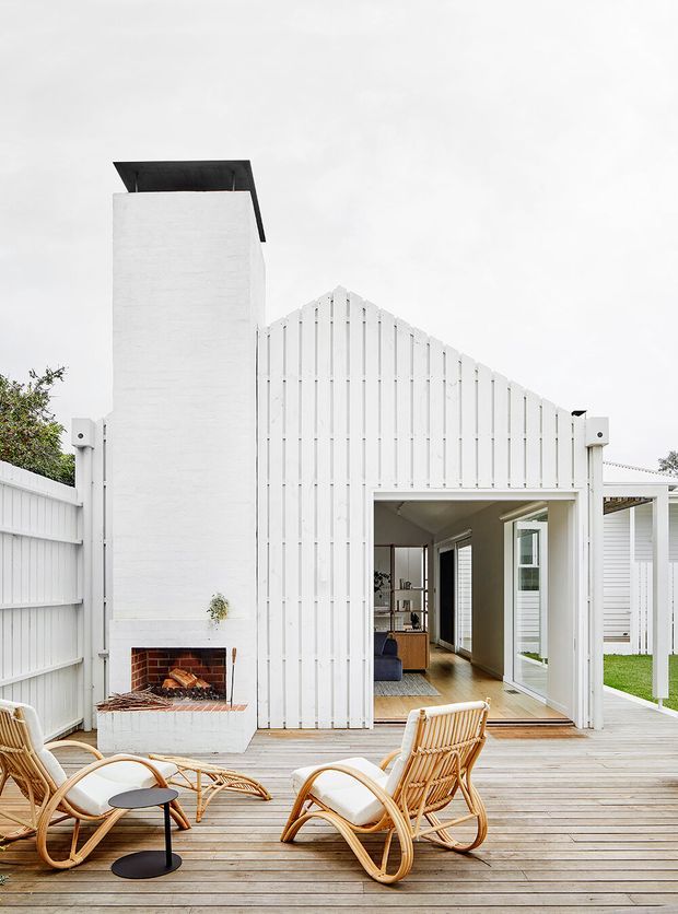 A bright white residential exterior centered on a gabled, vertically clad extension opening onto a timber deck. The facade includes a tall chimney with an outdoor brick fireplace, large glazed doors leading into a warm timber-floored interior, and a covered side section. Rattan lounge chairs and a small black table furnish the deck, creating a relaxed indoor-outdoor entertaining area.