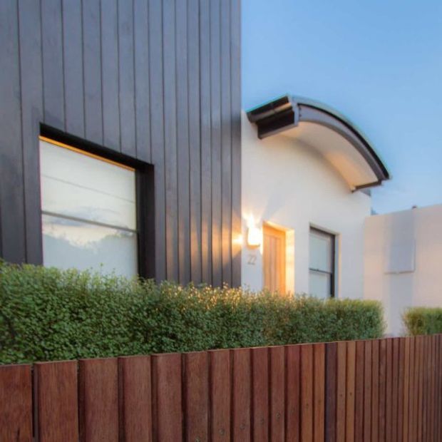 Exterior architectural view of a contemporary residence at dusk, featuring dark vertical cladding, a white rendered entry volume with a distinctive curved projecting roof, large windows, warm entry lighting, and a timber front fence with clipped hedging.