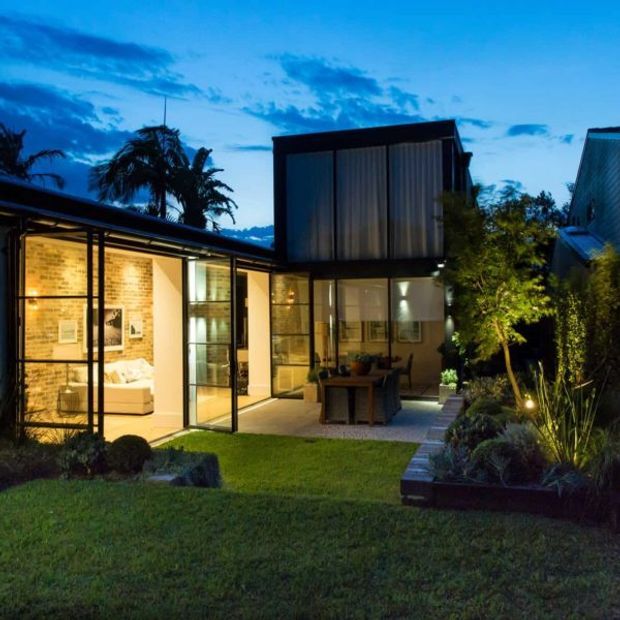Twilight exterior view of a contemporary residential extension with extensive floor-to-ceiling glazing, a dark-framed upper-level box volume, exposed brick interior walls, and a sheltered outdoor dining area opening onto a landscaped garden. Warm interior and landscape lighting contrast with the cool blue evening sky.
