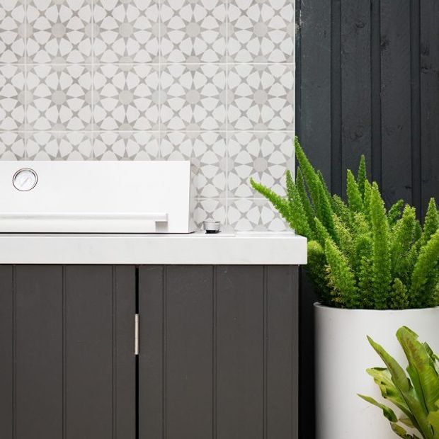 Close-up of a contemporary bathroom vanity featuring dark vertically grooved cabinetry, a white countertop and basin, and a patterned geometric grey-and-white tile splashback. Potted green plants add a natural accent beside the vanity.