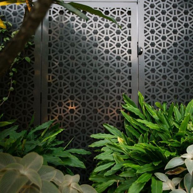 A close-up exterior view of a dark metal decorative privacy screen or gate with an intricate repeating geometric pattern, partially concealed by lush tropical foliage and broad-leaf planting. The screen has a narrow metal-framed door with a small round handle, creating a refined garden entry or boundary feature.