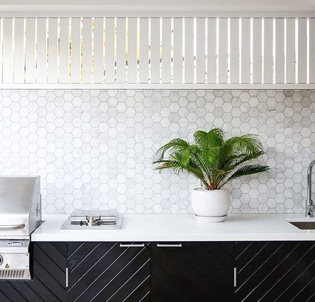 Bright contemporary kitchen vignette with a white benchtop, black diagonal-grain cabinetry, a white hexagonal tile splashback, stainless-steel appliances and fixtures, and a potted palm. White vertical blinds provide filtered daylight above the splashback.