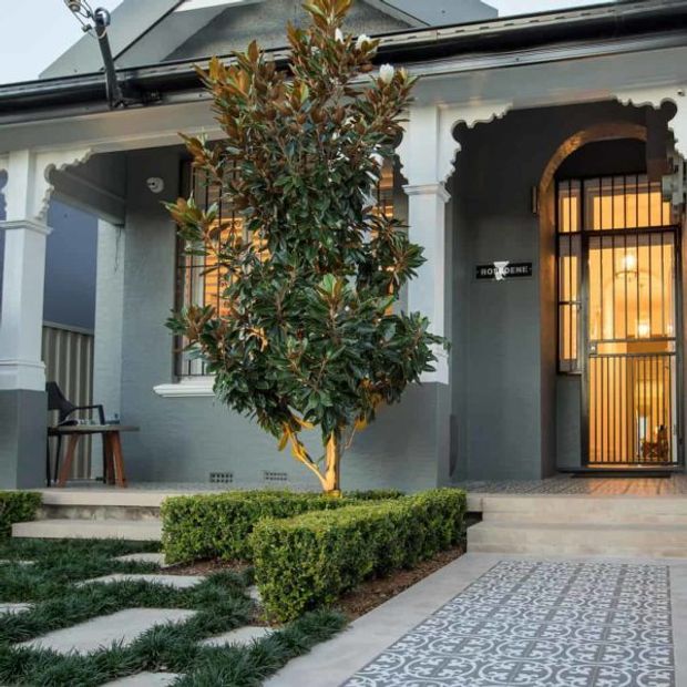 Street-facing heritage-style house entrance with a grey rendered facade, ornate white architraves and columns, barred glazed entry doors, tiled front steps, patterned pathway, clipped hedges, and a central ornamental tree.