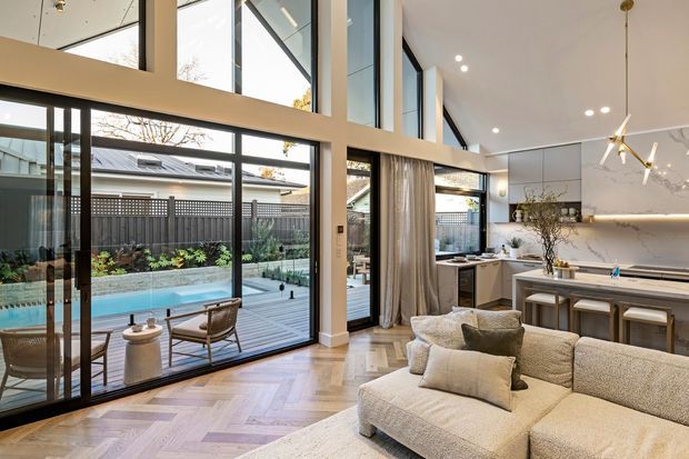 Open-plan contemporary living room and kitchen with a double-height glazed wall overlooking a backyard swimming pool and deck. The room features pale herringbone timber flooring, a large neutral upholstered sectional sofa, a minimalist kitchen with an island and stools, recessed lighting, and sculptural pendant lights. Black-framed floor-to-ceiling glazing creates a strong indoor-outdoor connection.