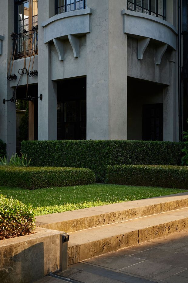 Exterior architectural view of a substantial rendered building with curved projecting balconies, decorative structural brackets, dark metal-framed windows, and ornate wrought-iron detailing. A formally landscaped frontage includes clipped hedges, lawn, raised concrete garden beds, and paved steps or walkways.