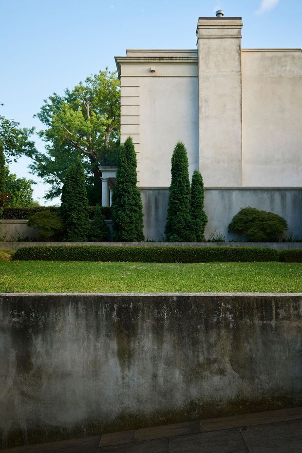Exterior architectural and landscape view showing a rendered, classically detailed building facade with a tall chimney, projecting cornice bands, and pilaster-like horizontal detailing. The facade is framed by clipped evergreen topiary, hedges, manicured lawn, and layered concrete retaining walls.