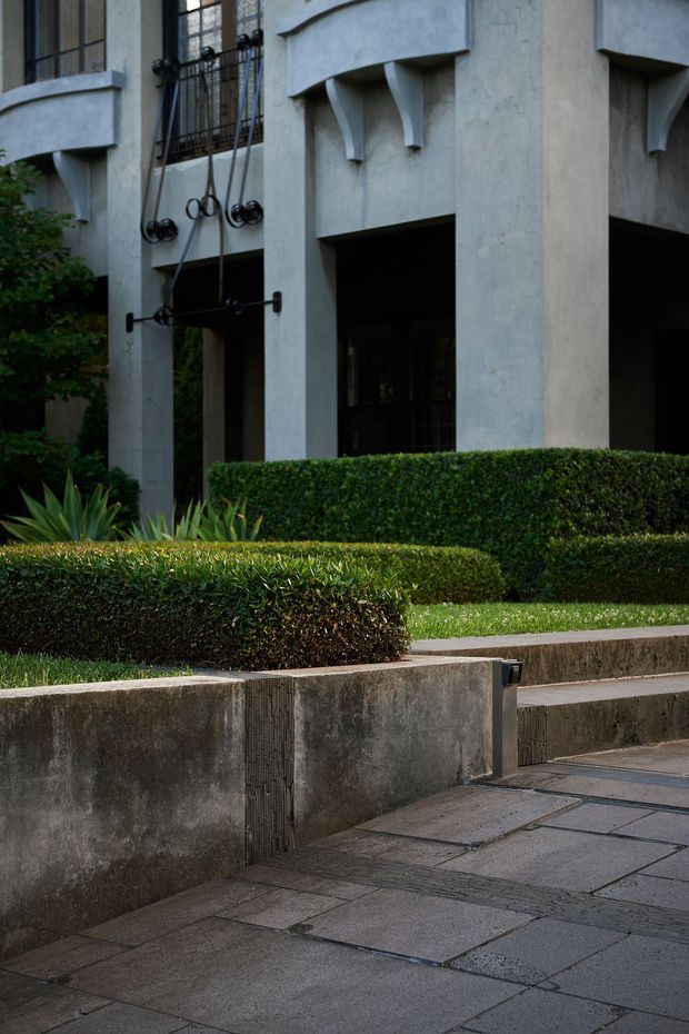 Exterior architectural photograph of a substantial rendered building with tall structural columns, curved projecting concrete balconies or ledges, decorative metalwork, and dark window openings. The foreground features formal clipped hedges, spiky ornamental planting, raised concrete garden beds, and a paved walkway.