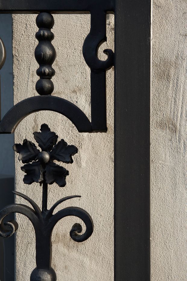 Close-up detail of an ornate black wrought-iron decorative grille or gate mounted against a light textured plaster wall. The metalwork features scrolls, turned spherical elements, and a central floral motif, with strong directional sunlight creating crisp shadows. No broader room or building context is visible.