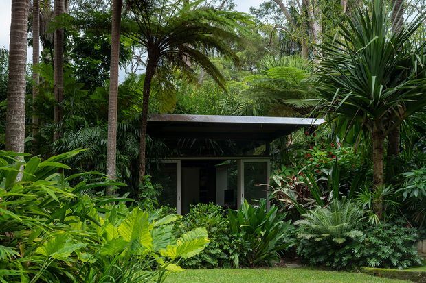 A small, low-profile pavilion or garden structure nestled within dense tropical planting. The building has a dark flat roof with broad overhangs, dark wall surfaces, and glazed doors or windows opening toward the lush garden. Tall palms, tree ferns, broad-leaved plants, and layered greenery dominate the setting.