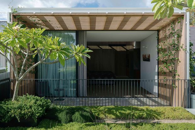 Contemporary residential facade with a covered balcony or terrace, vertical timber screening, a broad glazed opening, and a flat projecting pergola roof. Lush planting and a simple dark metal balustrade frame the outdoor space.