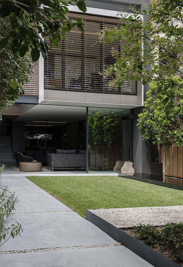A contemporary two-storey residence viewed from a landscaped front courtyard. The dark rendered facade has extensive floor-to-ceiling glazing, a projecting upper level with horizontal timber screening, and a recessed ground-floor living area opening onto the garden. Broad concrete-paver paths, a clipped lawn, gravel beds, raised dark garden edging, timber fencing, and established planting create a restrained modern landscape.