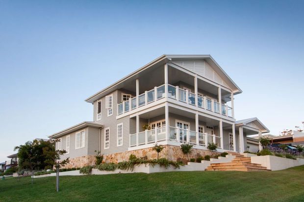 Exterior view of a large elevated coastal-style residential house with grey weatherboard cladding, extensive white-trimmed glazing, multiple covered balconies, a pitched gabled roof, and landscaped stepped grounds.
