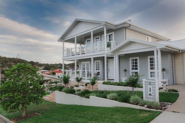 Exterior view of a light-colored two-storey suburban home with a prominent covered upper balcony, white railings, pitched gable roof sections, and horizontal weatherboard cladding. The landscaped front yard includes lawn, raised garden beds, shrubs, and a concrete pathway. The surrounding residential hillside is visible in the background.