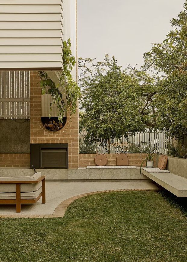 A landscaped residential courtyard with a contemporary outdoor entertaining area. The space features a built-in concrete bench with timber-framed cushions, a freestanding timber outdoor chair, a brick garden wall, curved brick paving edging, lawn, mature trees, and a compact dark outdoor fireplace or barbecue integrated into the brick facade. The building has pale horizontal cladding and vertical screening elements, creating a warm, refined outdoor setting.