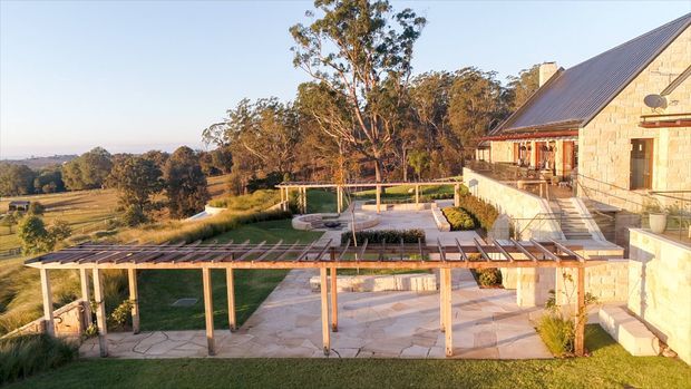 A rural stone-clad residence viewed from above, with a steep dark metal pitched roof, broad paved terrace, timber pergola structures, outdoor seating areas, lawns and landscaped grounds overlooking a wooded and agricultural valley.