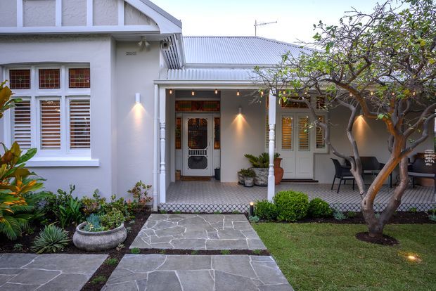 Front exterior of a compact heritage-style home, featuring pale rendered walls, a corrugated metal roof, decorative timber-framed windows with shutters, a glazed entry door, and a covered verandah. A paved stone path leads through a landscaped front garden with established trees, planting beds, and a small seating area.