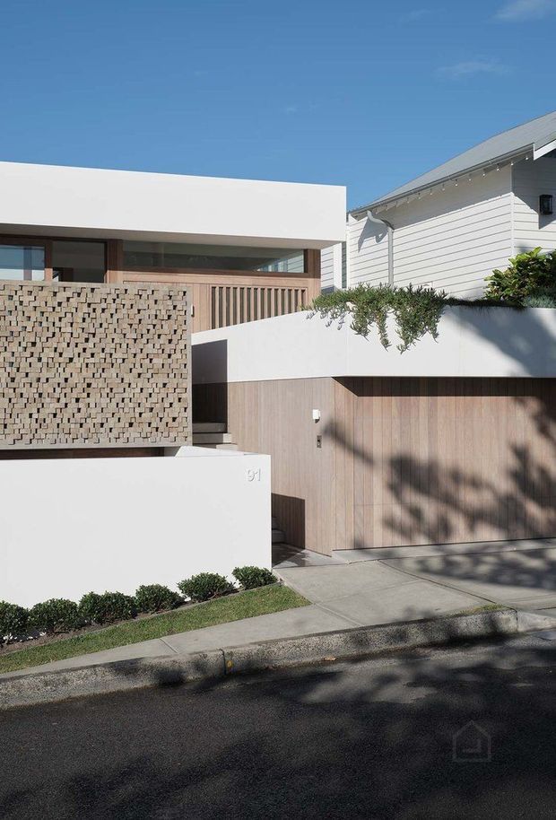 Street-facing view of a contemporary residential façade combining crisp white rendered volumes, a textured breeze-block privacy screen, vertical timber cladding, a timber garage door, and a low planted terrace wall. A neighbouring weatherboard house is visible at right. The frontage includes a concrete footpath, landscaped shrubs, and strong sunlight with tree shadows.