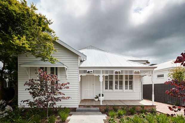 Front exterior of a renovated single-storey timber-clad home with a white corrugated metal roof, traditional gabled form, and a prominent covered verandah featuring decorative timber fretwork and posts. White-framed windows with shutters, brick steps, a paved entry path, and landscaped garden beds are visible beneath a dramatic cloudy sky.
