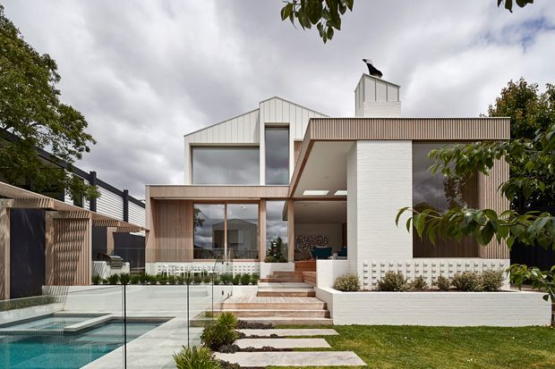 Contemporary two-storey residential facade viewed from a landscaped backyard, featuring white rendered walls, vertical timber-look cladding, extensive glazing, a projecting flat roof, a rooftop chimney, and a swimming pool with frameless glass fencing in the foreground. Stepped stone landscaping and a patterned breeze-block wall complement the clean geometric design.