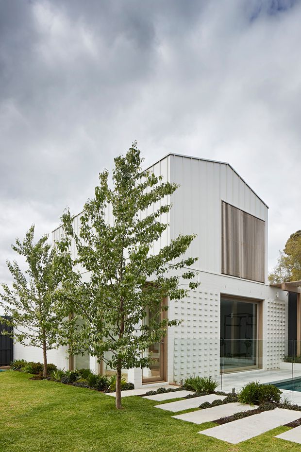 A contemporary multi-level house exterior with a tall gabled volume clad in white vertical metal panels, a large screened upper opening, textured white masonry at ground level, extensive glazing, and timber-framed doors. A landscaped lawn, young trees, stepping-stone path, and a partially visible swimming pool surround the building.