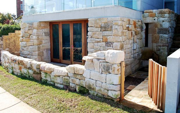 Exterior view of a contemporary multi-level building with a prominent lower facade constructed from rough-cut pale natural stone. Timber-framed glazed double doors open onto a small enclosed stone terrace, with stone retaining walls and steps leading around the side. A glass-balustraded upper level and modern rendered elements contrast with the masonry base.