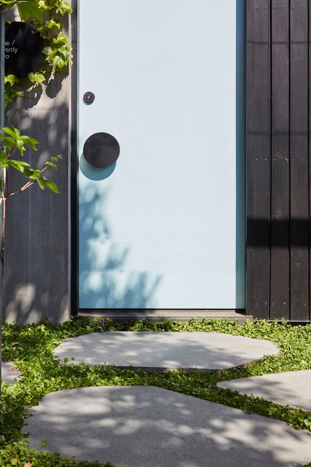 Close-up exterior view of a minimalist pale blue entry door with a large matte black circular handle and small circular lock, set within dark vertical timber cladding. Irregular concrete stepping stones lead to the entrance through low green groundcover, with leafy shadows and climbing vegetation adding natural texture.